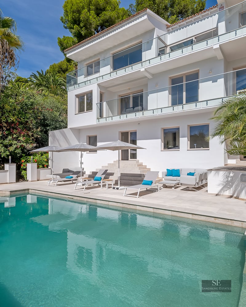 Three-story white villa featuring a large turquoise pool, sun loungers, and palm trees under a clear blue sky.