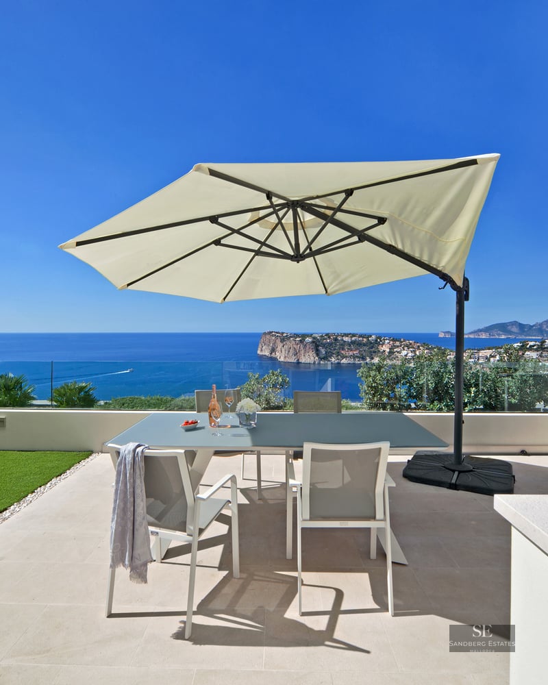 Outdoor terrace with dining table, sun umbrella, BBQ grill, and stunning ocean views under a clear blue sky.