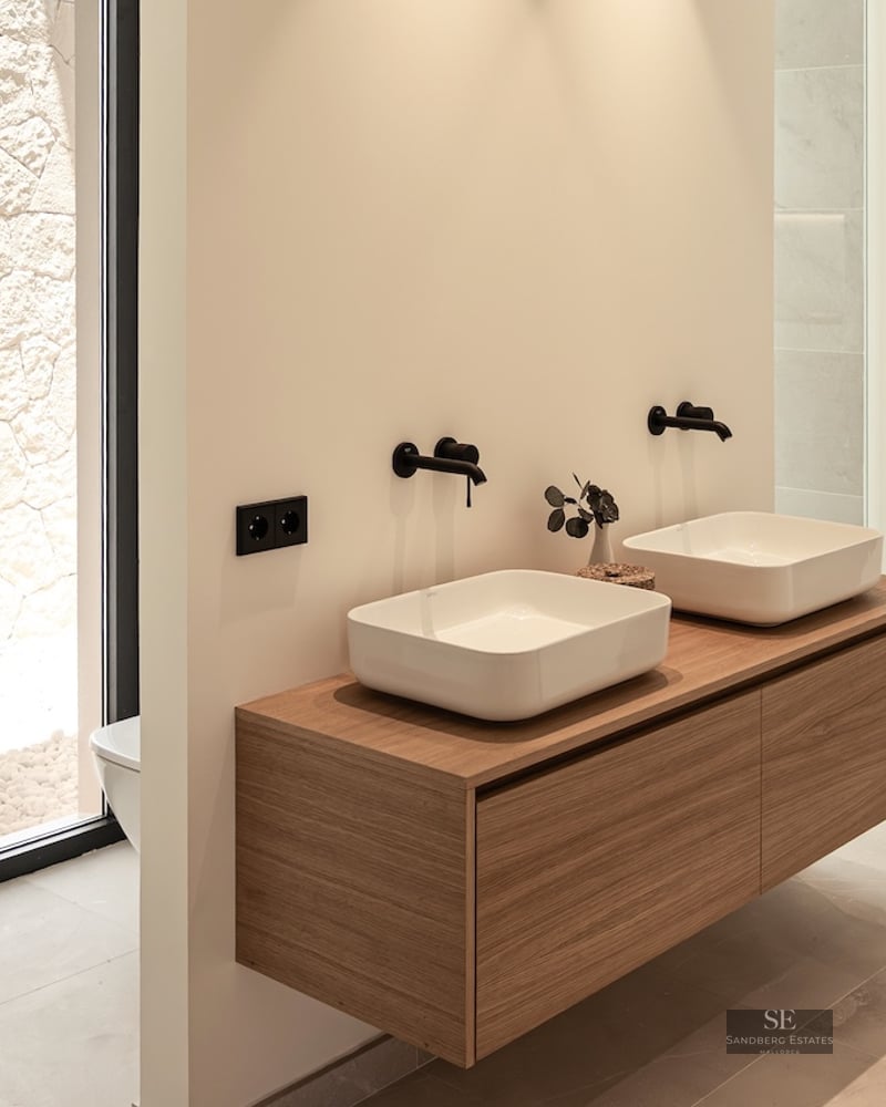 Two white vessel sinks on a floating wood vanity with matte black wall faucets in a bright modern bathroom.
