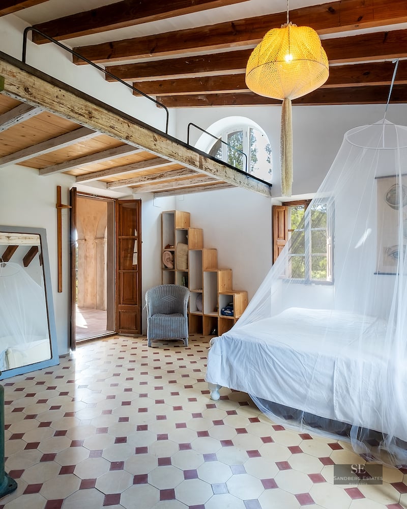 Spacious bedroom with white bed, mosquito net, wooden mezzanine loft, exposed beams, and decorative tiled floor.