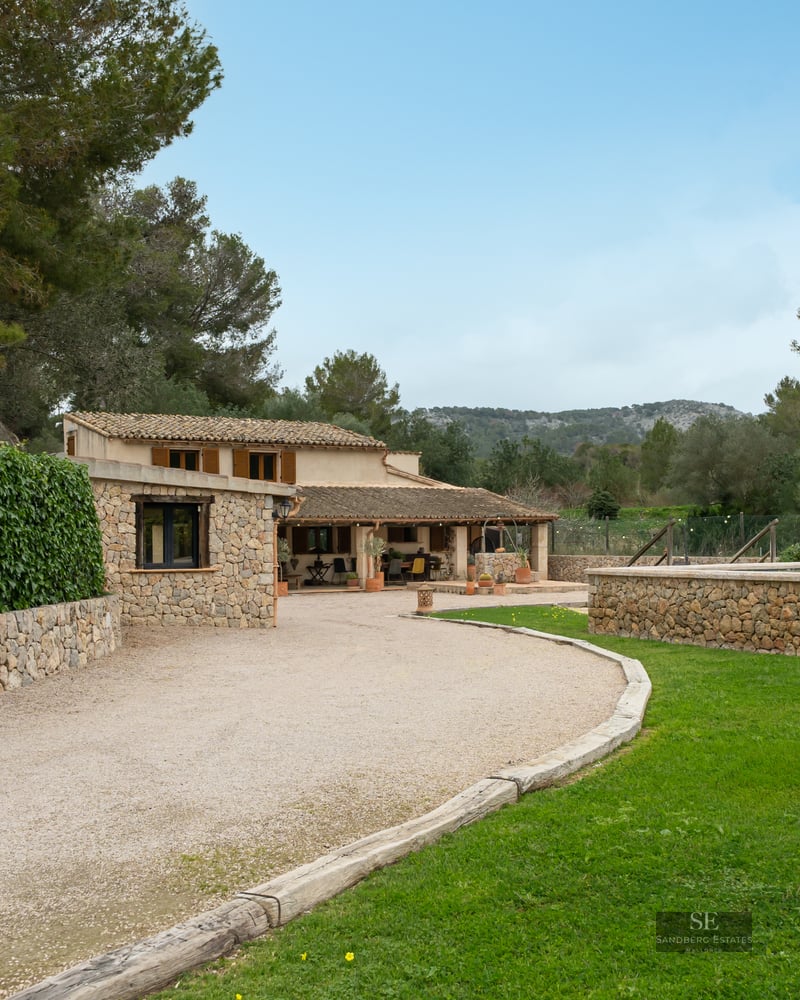 A rustic stone house with a gravel driveway, lush green lawn, and an elevated stone pool area under a clear sky.