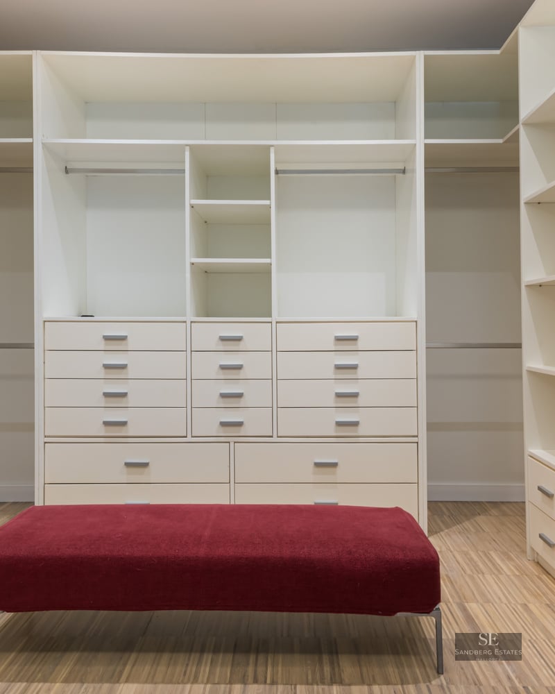 Spacious white walk-in closet with numerous shelves, drawers, wooden flooring, and a central red velvet ottoman.