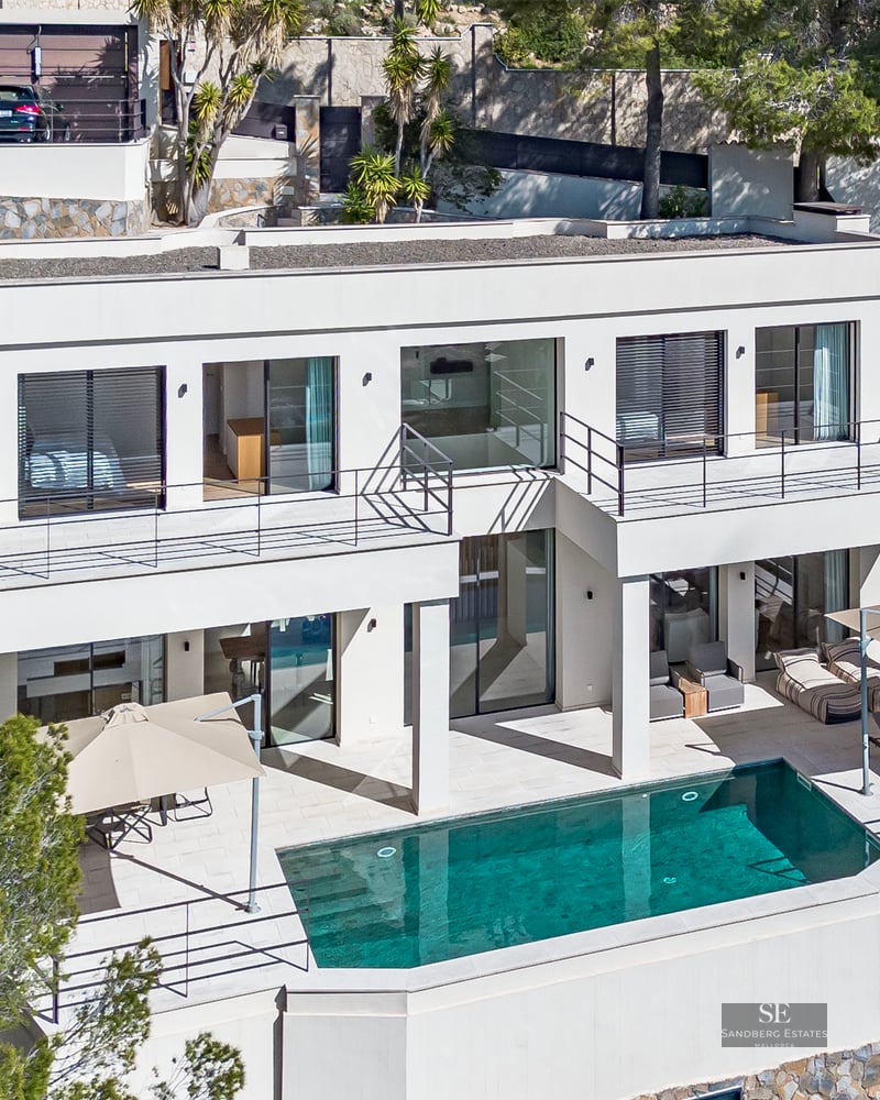 Aerial view of a white modern villa with a turquoise pool, sun loungers, and pine trees on a hillside.