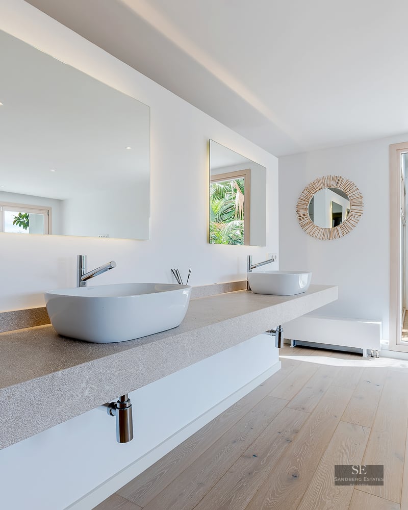 Bright bathroom with stone floating vanity, two white sinks, large mirrors, and a window to a tropical garden.