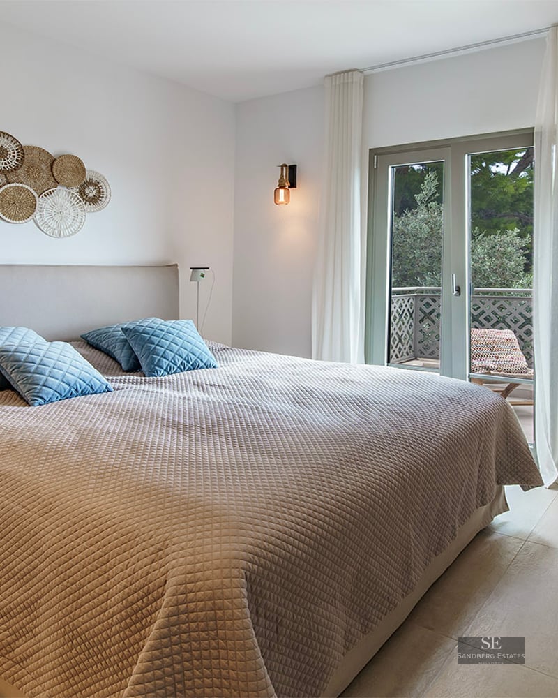 Modern bedroom featuring a large bed, beige textured bedding, blue pillows, wooden desk, and glass door to a balcony.
