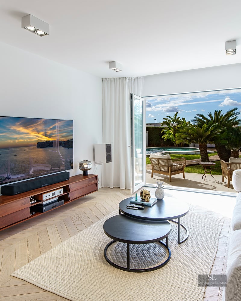 Bright living room with white sofa, herringbone wood floor, and open doors to a sunny terrace with palms.