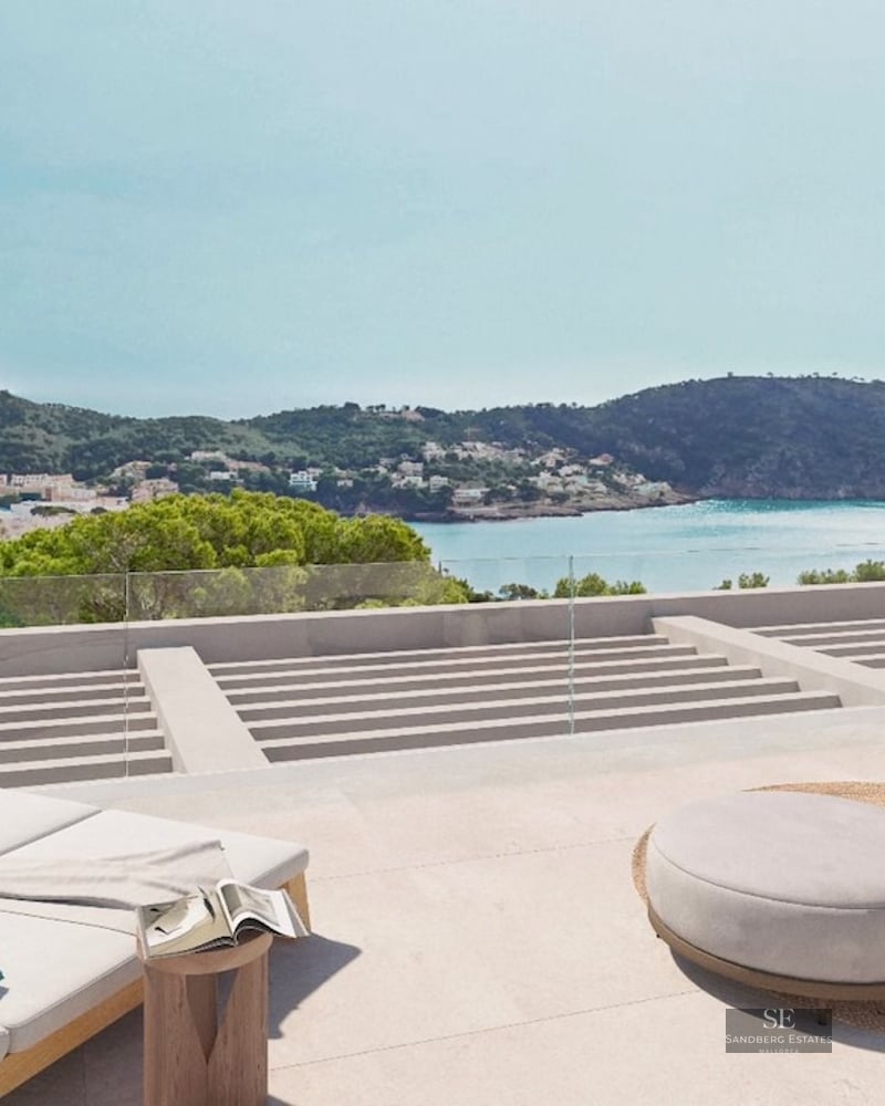 Modern stone terrace with sun loungers and designer furniture overlooking a Mediterranean bay and green hills.