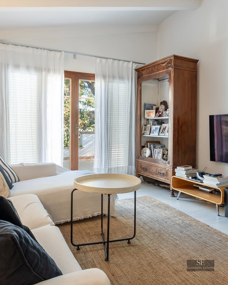 Bright living room with white sofas, a large wooden armoire, wall-mounted TV, and natural light from a terrace door.