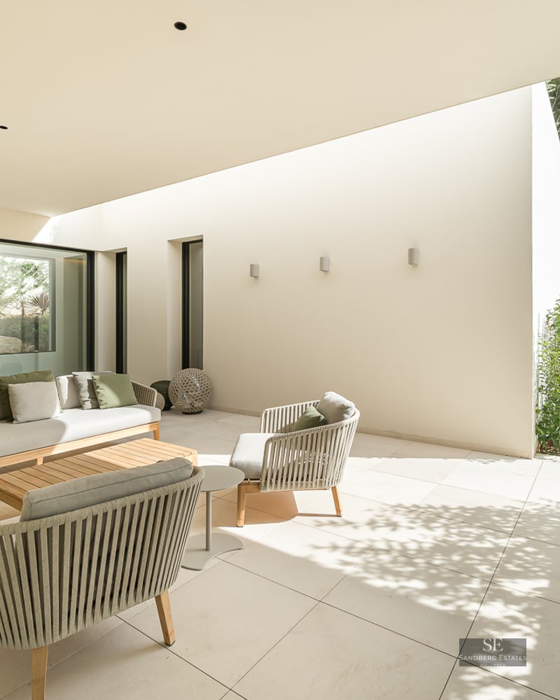 Luxury outdoor terrace with a beige sofa, green cushions, and woven chairs on a stone patio overlooking a garden.
