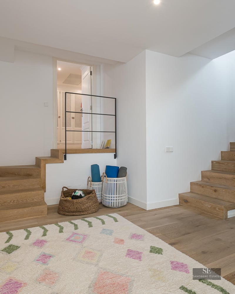 Pièce blanche avec deux escaliers en bois, fresque colorée et tapis de yoga dans des paniers.