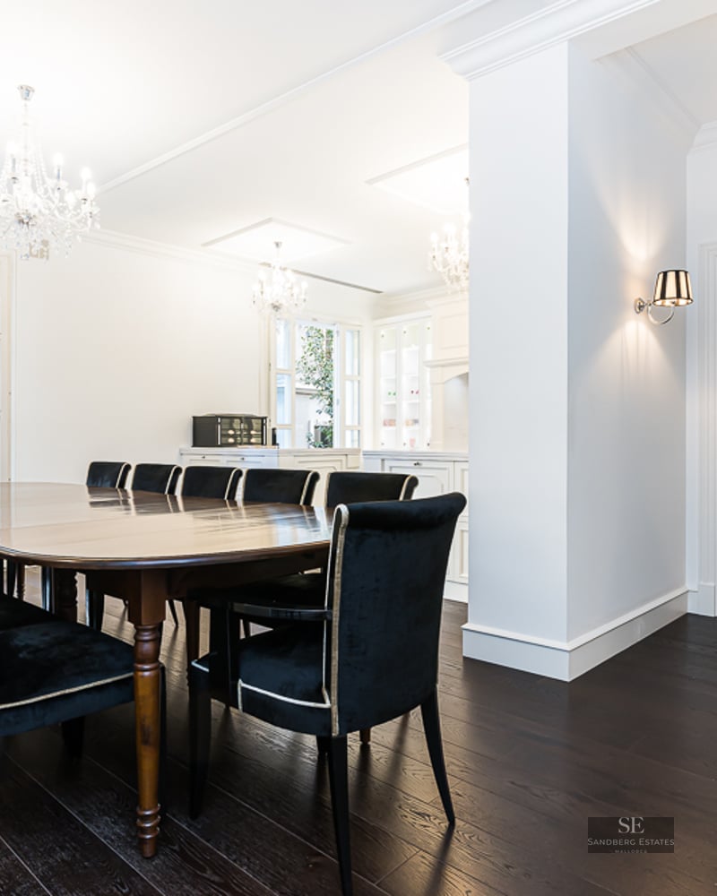 A large dark wood dining table with black velvet chairs under crystal chandeliers in a bright white room.