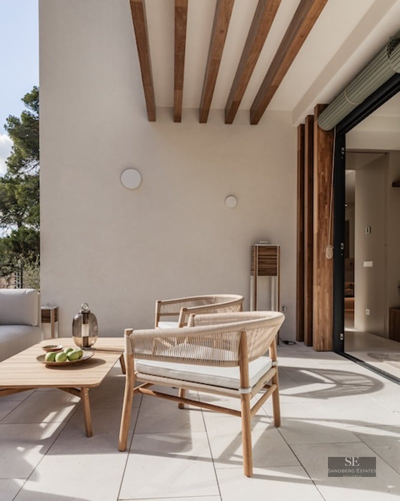A sunlit modern terrace with a white sofa, wooden furniture, and views of lush green trees through glass doors.