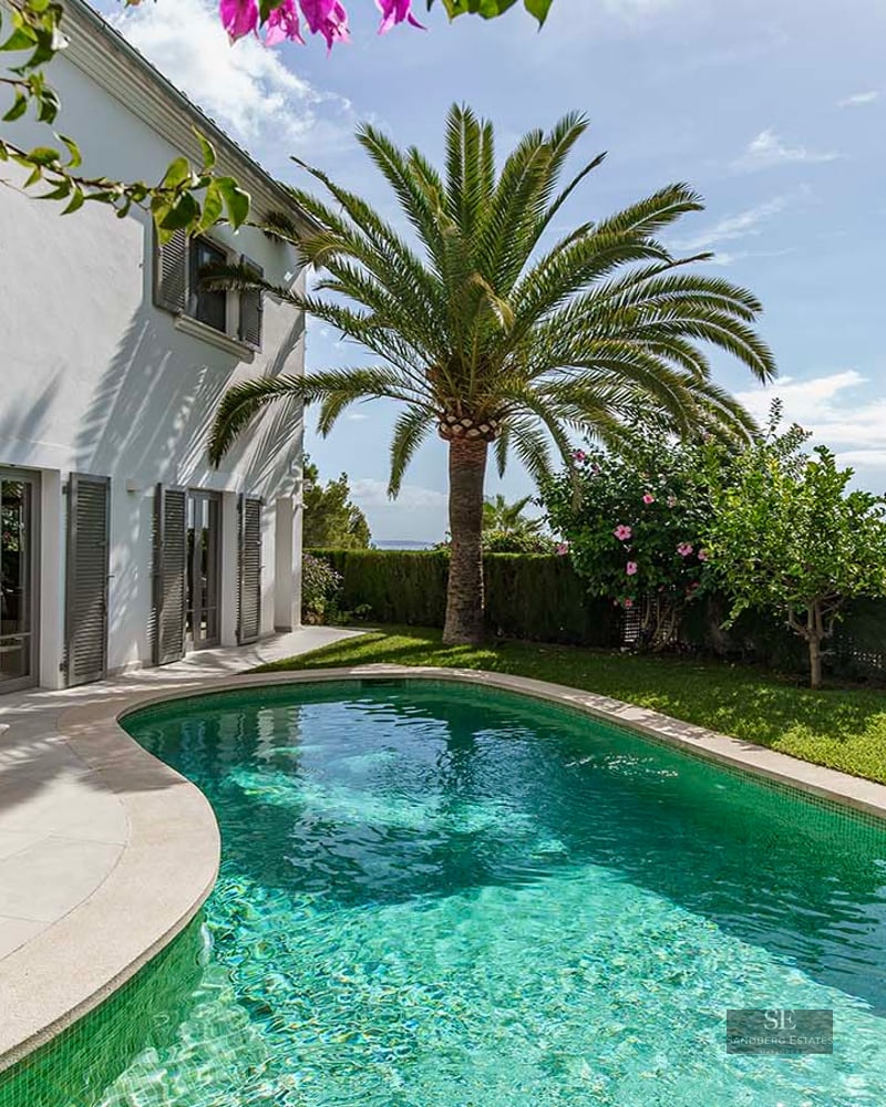 A curved turquoise pool bordered by a lush lawn, a palm tree, and a white villa with grey shutters under a blue sky.