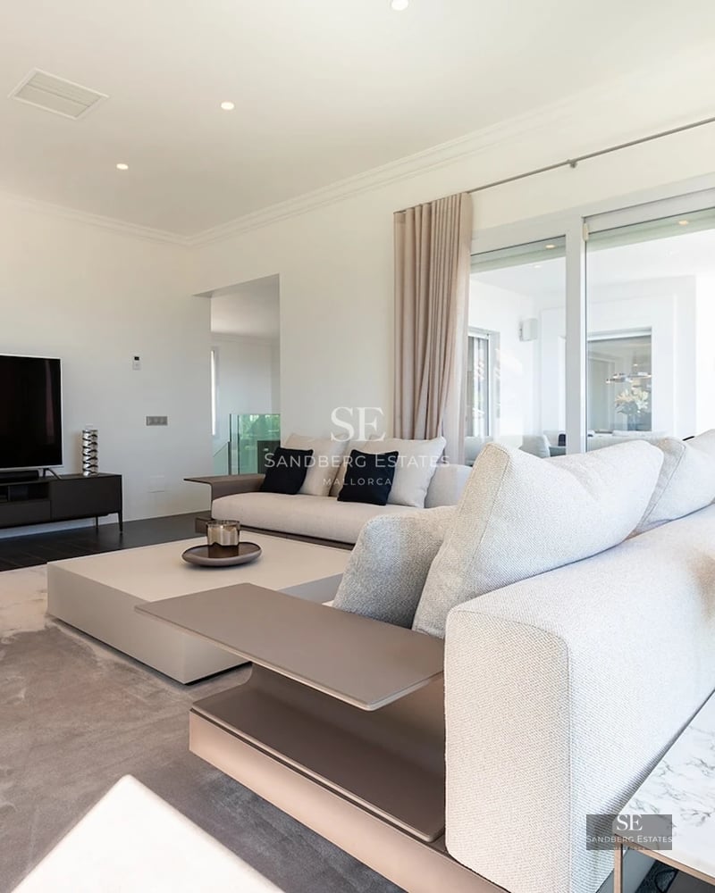 Modern living room with large grey sofas, coffee table, big screen TV, and floor-to-ceiling glass doors.
