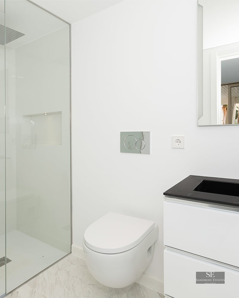 A bright, minimalist bathroom featuring a glass walk-in shower, a white wall-hung toilet, and a black and white vanity.