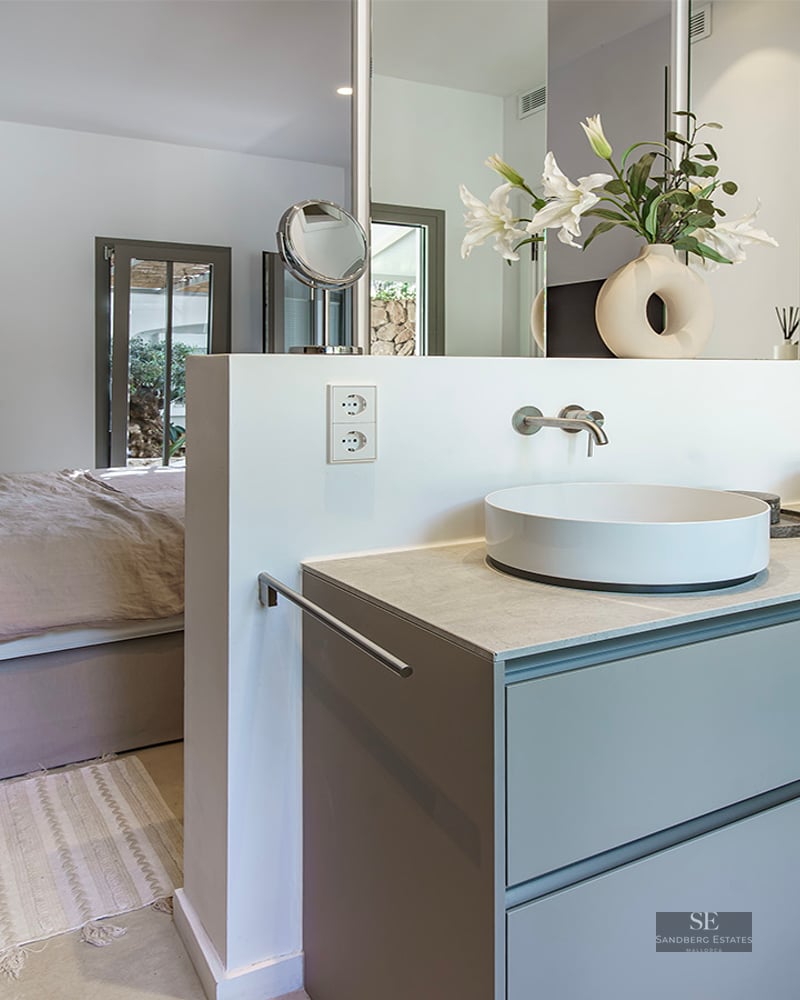 Open-plan master bedroom featuring a double vanity with two vessel sinks and a large bed with blue pillows.