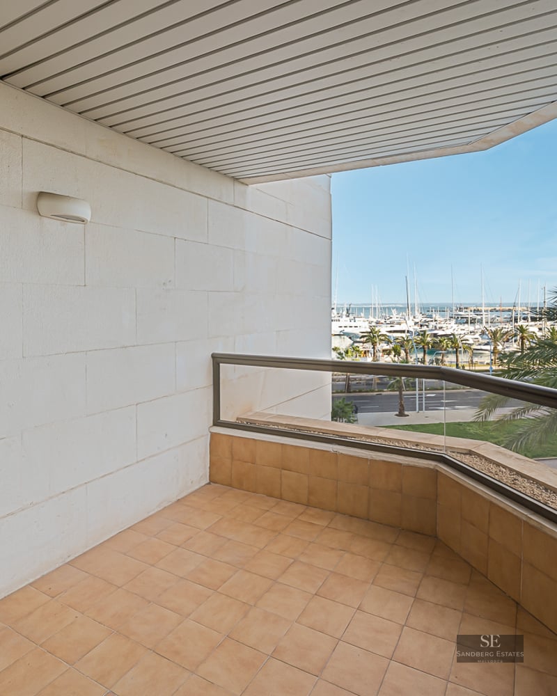 Covered terrace with glass railing overlooking a harbor with yachts and palm trees under a clear blue sky.