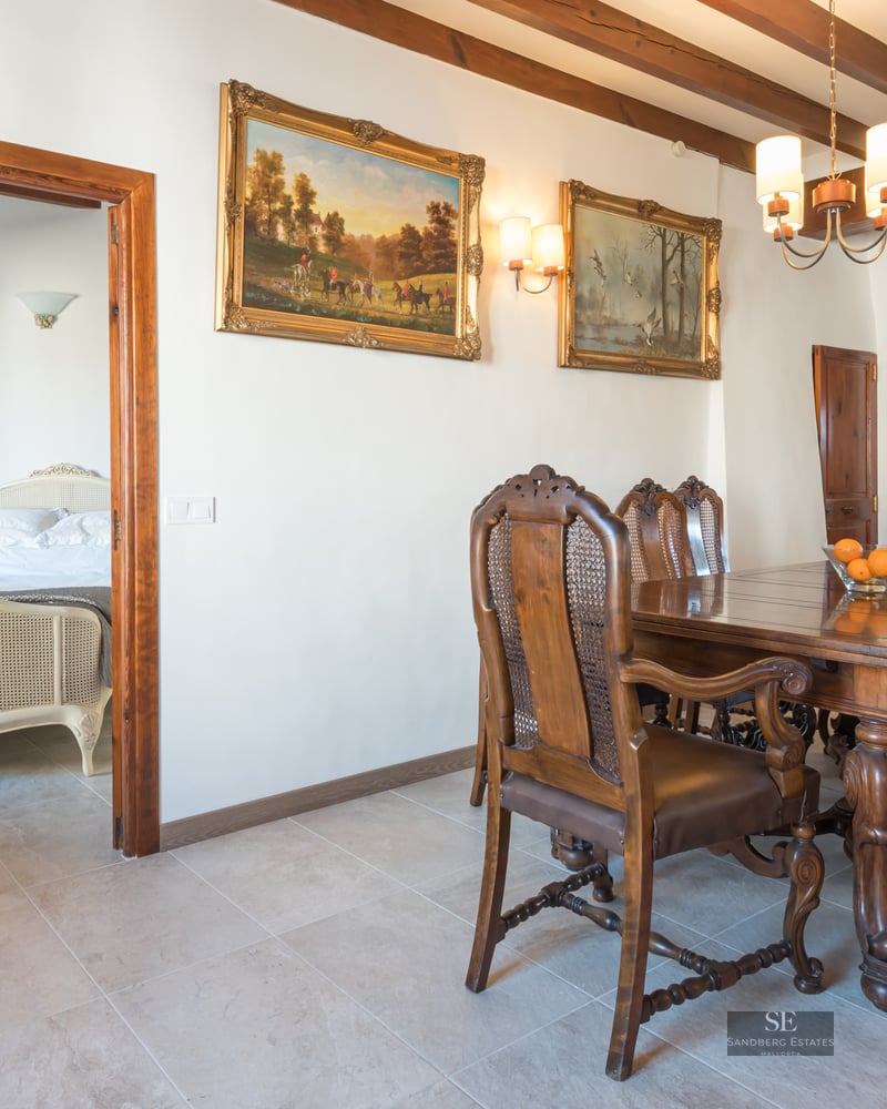 Dining room featuring a carved wood table, leather and cane chairs, beamed ceilings, and a view into a bedroom.