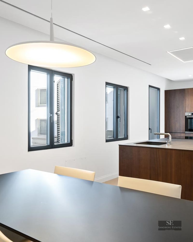 Open-plan kitchen featuring a dark wood island, black dining table, white walls, and three rectangular windows.