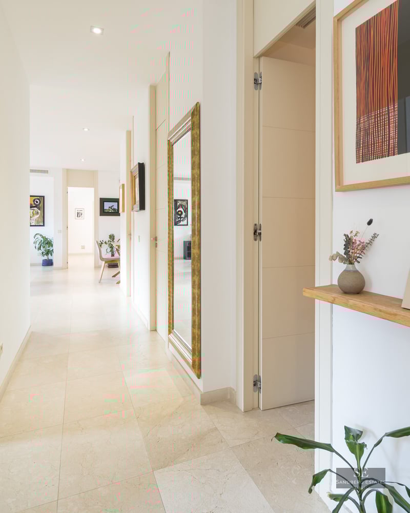 Long white hallway with stone floors, a large gold-framed mirror, and colorful art pieces.