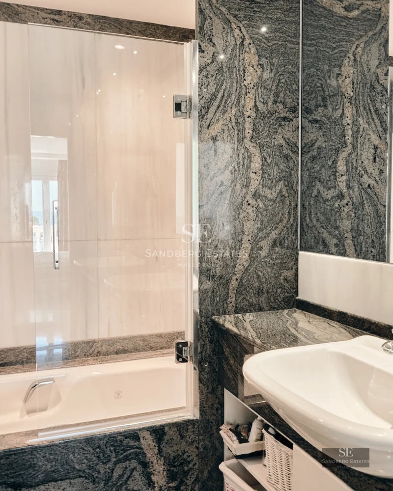 Bright, spacious living room with large windows and balcony overlooking the sea. Light wood floors and modern decor.