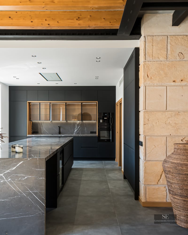 A contemporary kitchen featuring a grey marble island, black cabinetry, exposed wooden ceiling beams, and a stone wall.