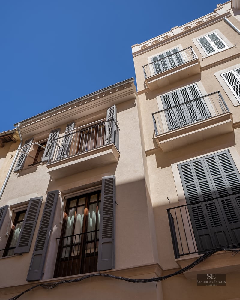 Low angle view of a beige building facade with green shutters and wrought iron balconies against a clear blue sky.