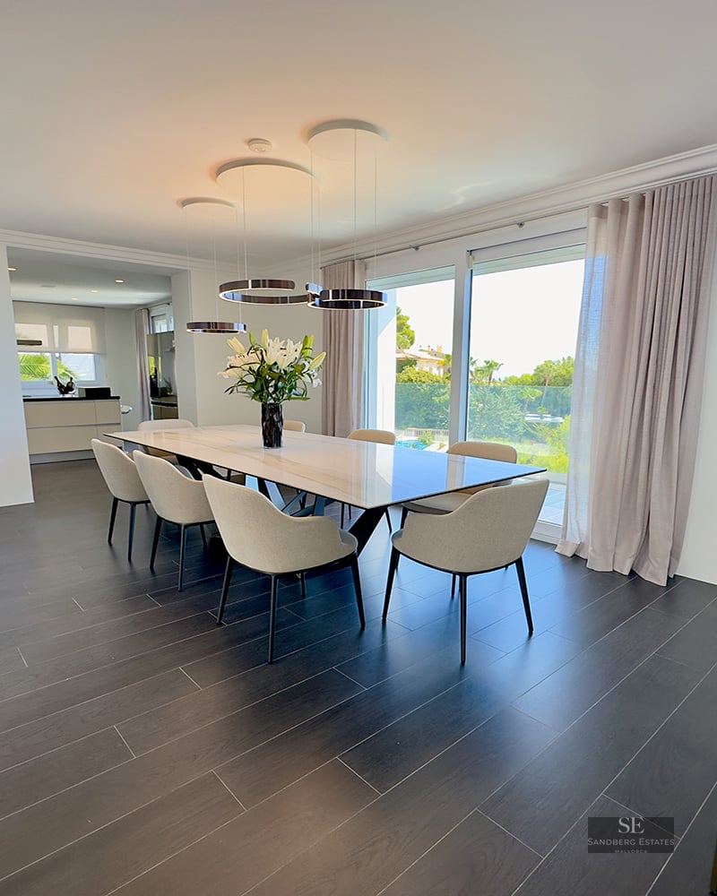 Minimalist dining room featuring a marble table, six beige chairs, dark flooring, and large windows viewing a garden.