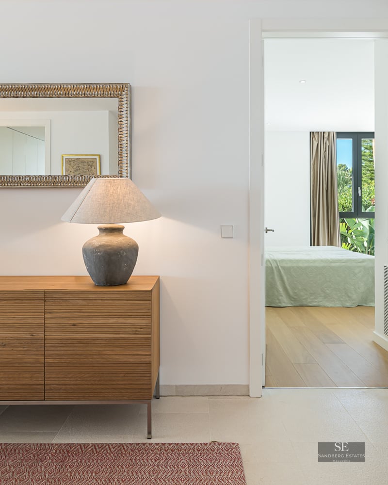 A wooden sideboard with two lamps and an ornate mirror in a bright hallway leading to a bedroom.