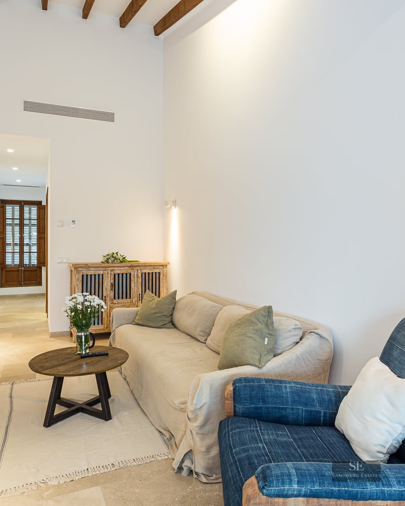 Bright living room with white walls, wooden ceiling beams, beige sofa, blue armchair, and round coffee table.
