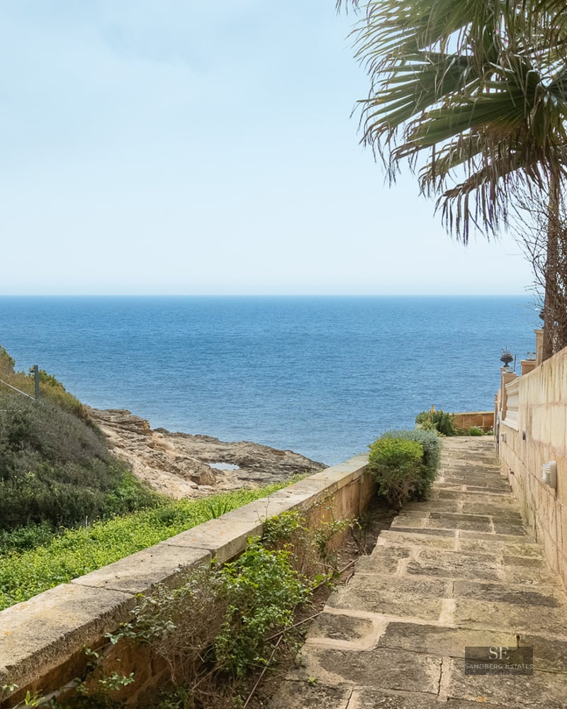 A stone pathway between a wall and greenery leads directly towards a vast, blue ocean.