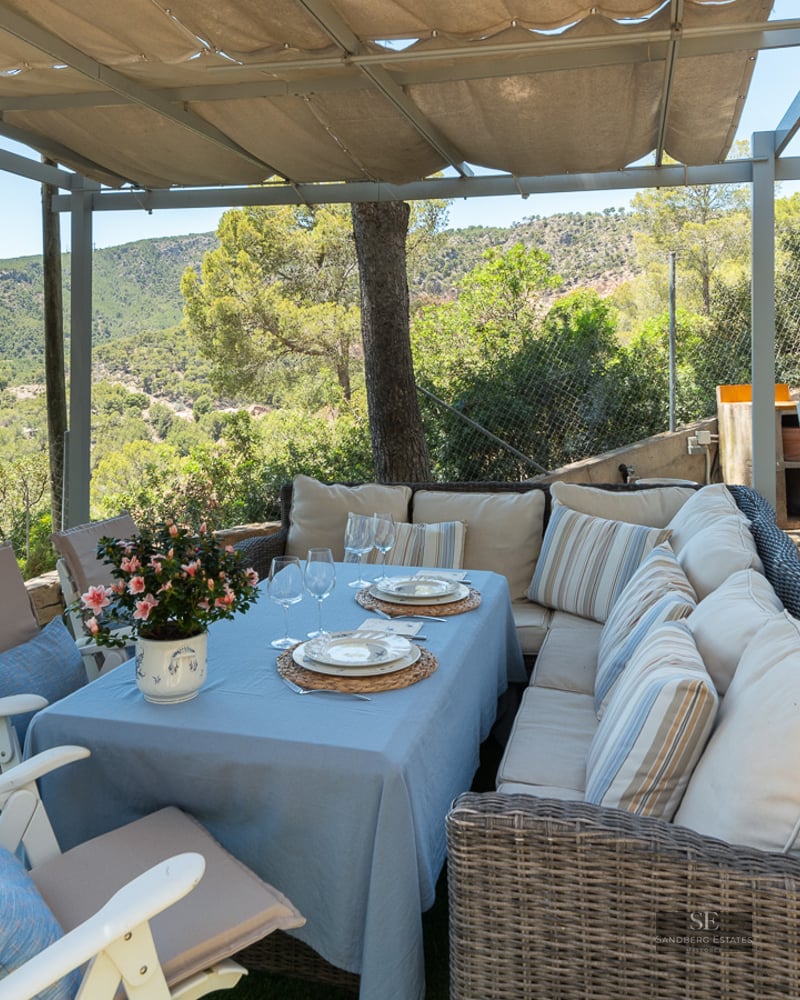 Outdoor dining area under a pergola with wicker sofas and views of green forested hills.