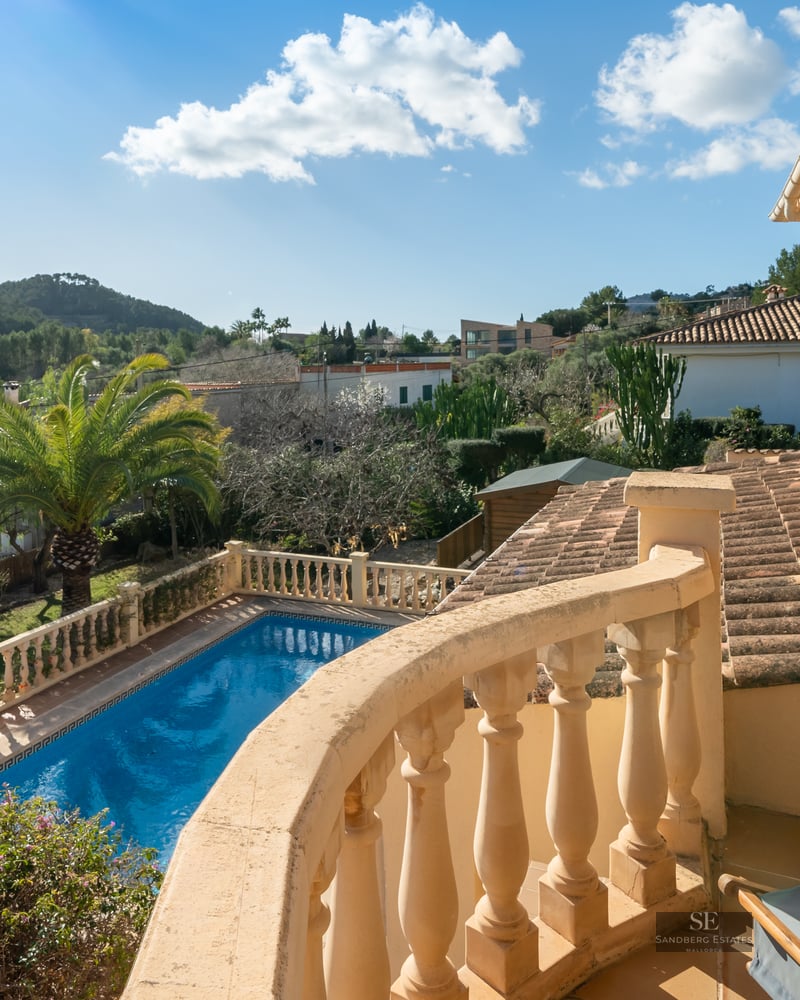 View from a stone balcony overlooking a blue swimming pool, lush gardens, and rolling hills under a clear blue sky.