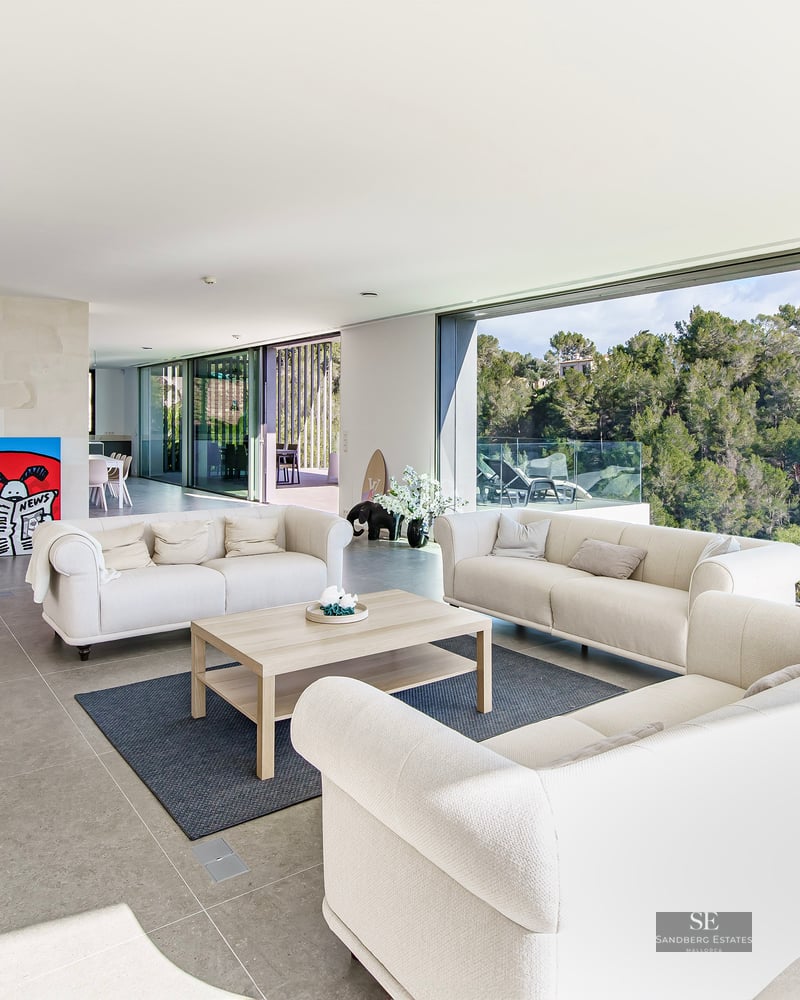 Spacious modern living room with cream sofas and large glass walls overlooking a lush green forest.
