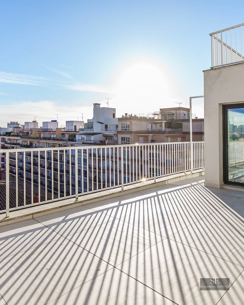 Spacious penthouse terrace with white tiles and railings, overlooking a city skyline under a clear, bright sun.
