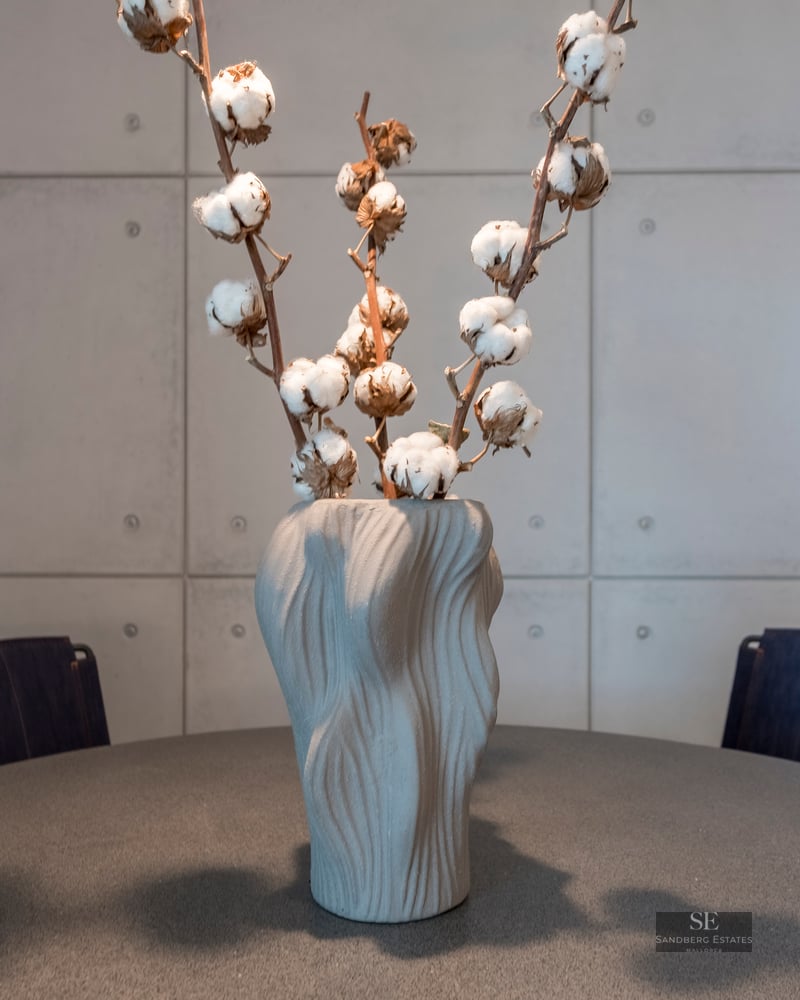 Textured gray vase with cotton branches on a dark table in front of a concrete panel wall.