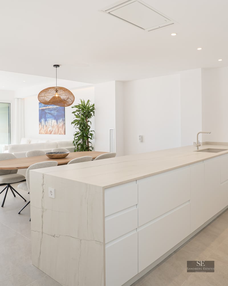Modern white kitchen island and wooden dining table in a bright open-plan apartment with terrace and city views.