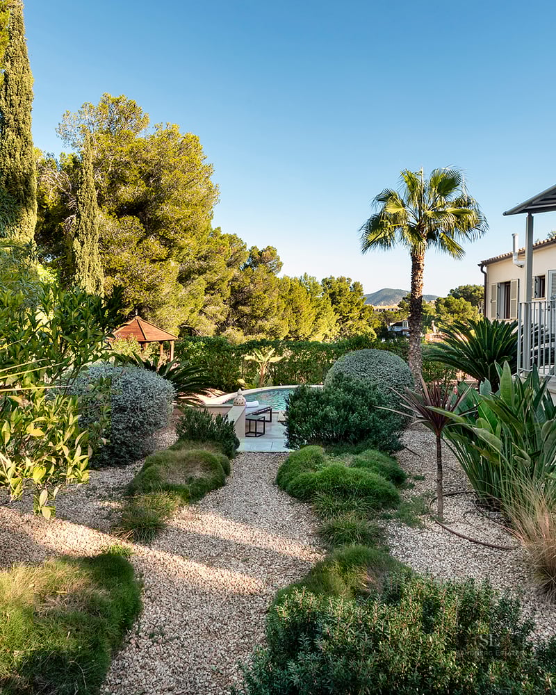 Lush landscaped garden with gravel paths leading to a swimming pool, next to a Mediterranean style villa.