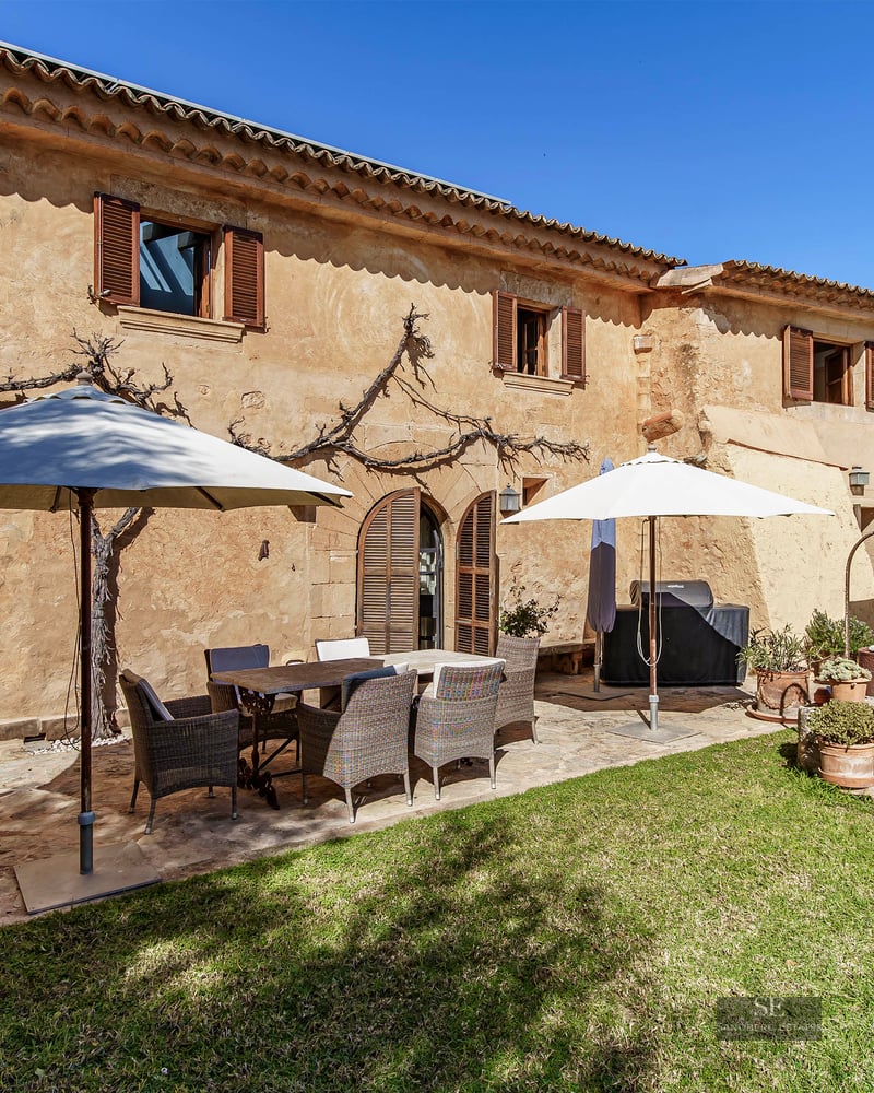 Sunny stone terrace with a dining table, wicker chairs, and white umbrellas next to a green lawn.