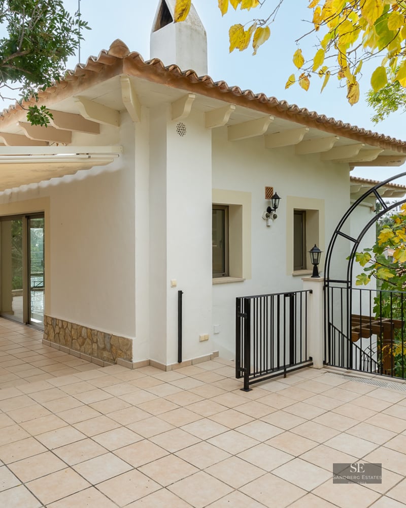 Spacious tiled terrace with a wrought iron dining set, white villa walls, and lush greenery under a retractable awning.