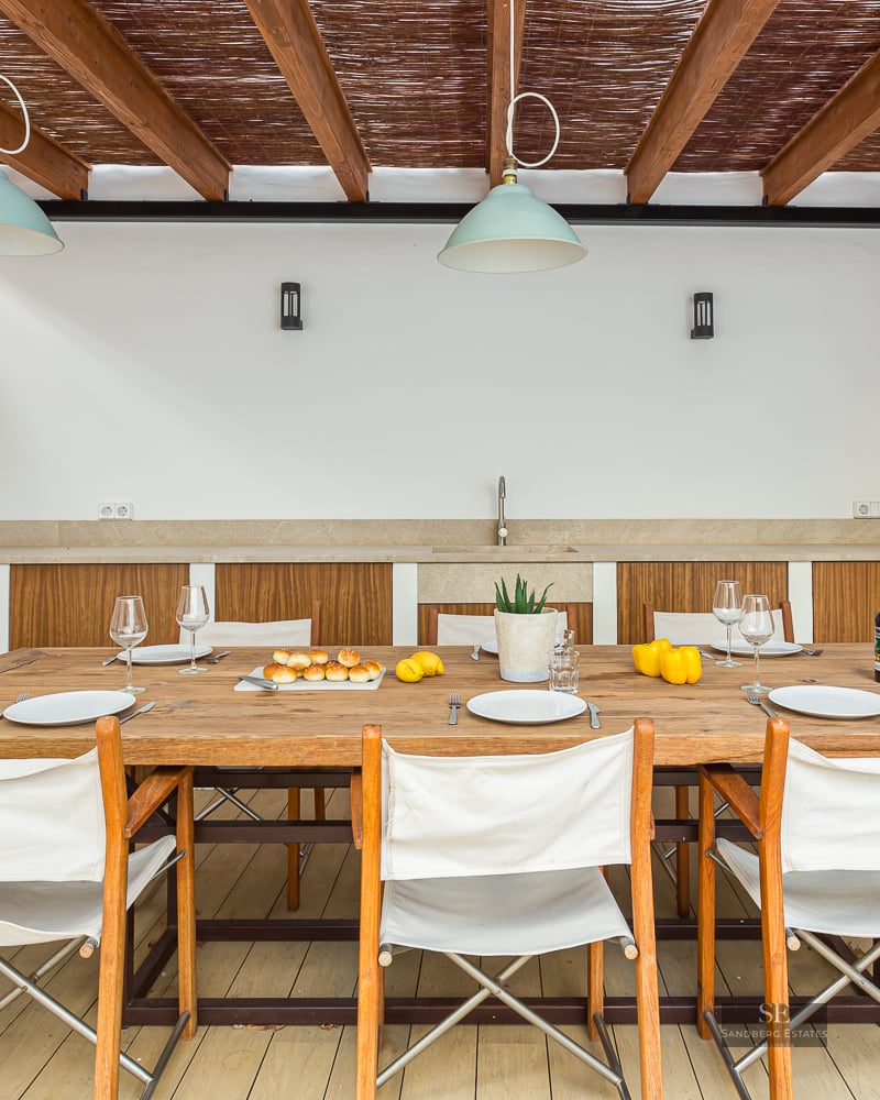 Covered terrace with large wooden table, white canvas chairs, and an outdoor kitchenette under a wooden pergola.