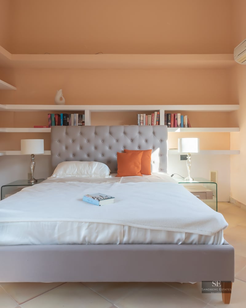 Bright bedroom featuring a tufted grey headboard, white bedding, built-in shelving, and terracotta tile floors.
