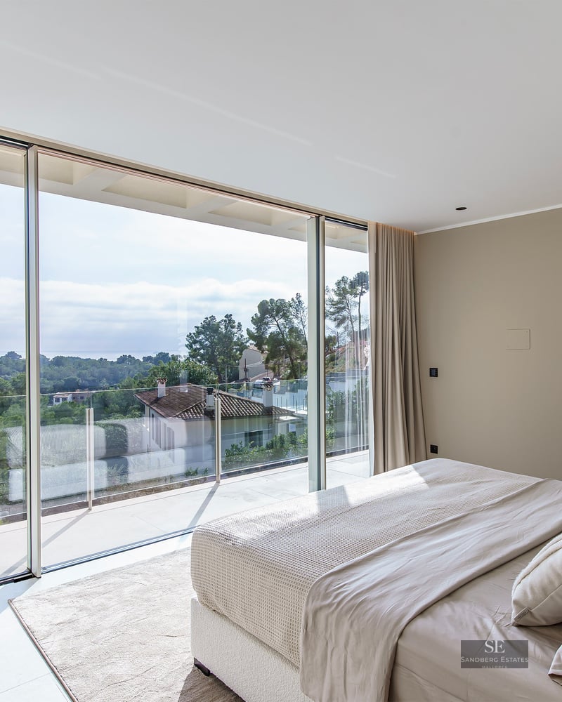 Luxury bedroom with floor-to-ceiling glass doors overlooking trees and houses, featuring a plush bed and boucle chair.