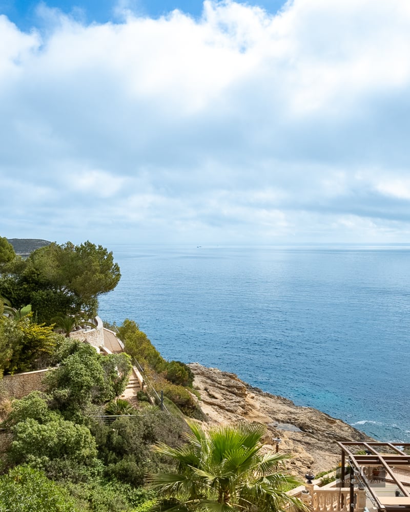Elevated view of a rocky coastline with deep blue sea, green pine trees, and a Mediterranean villa.