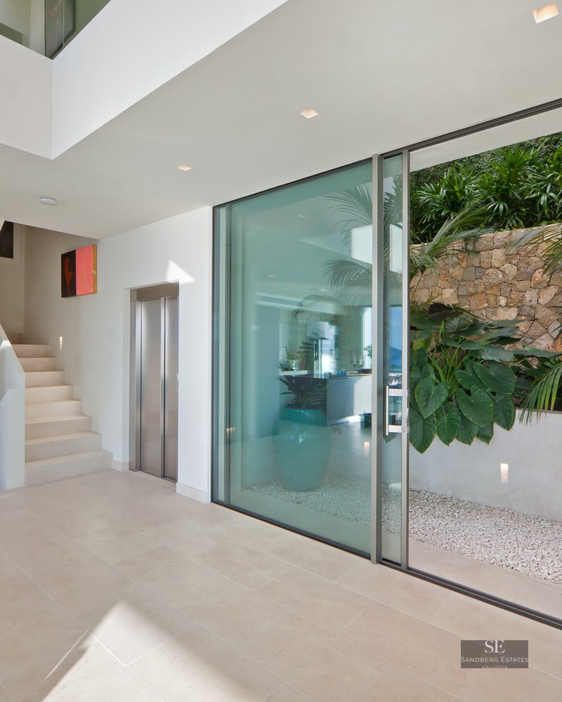 Minimalist lobby with light stone floors, an elevator, white stairs, and a glass door to a lush tropical patio.