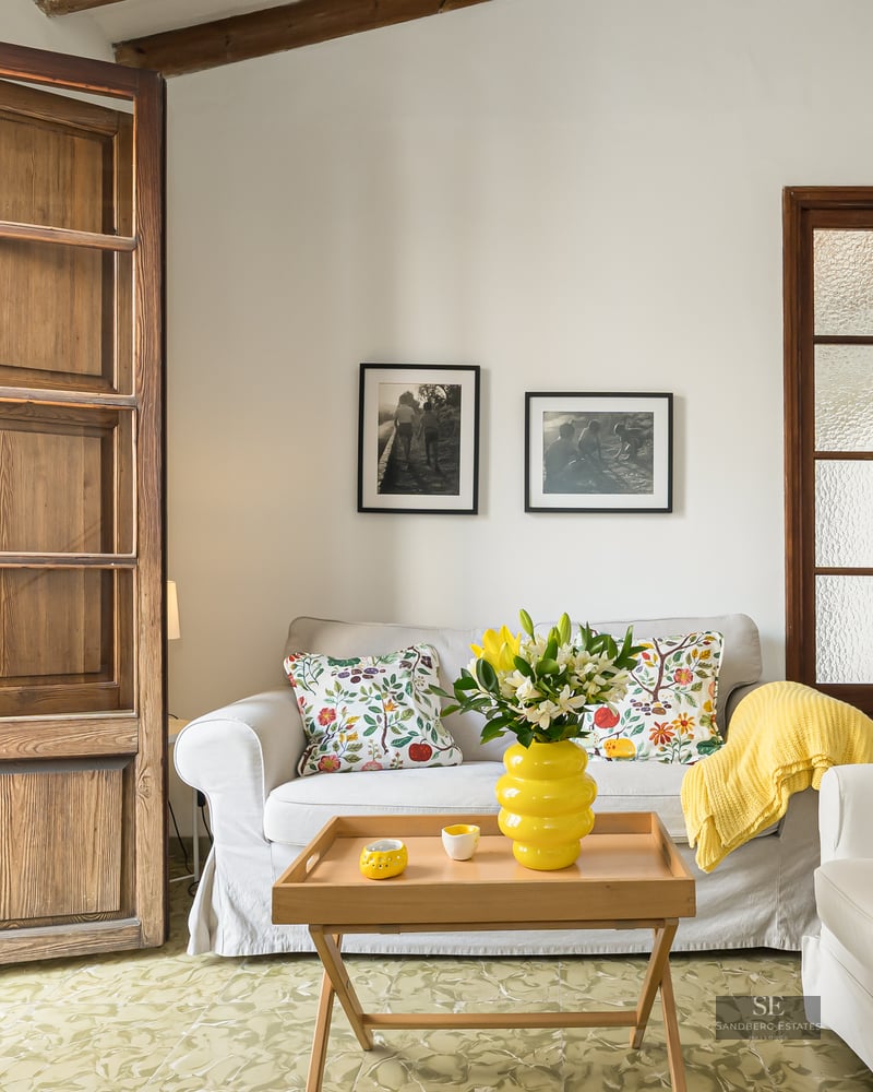Bright living room with white sofas, colorful cushions, and large wooden balcony doors opening to a city view.