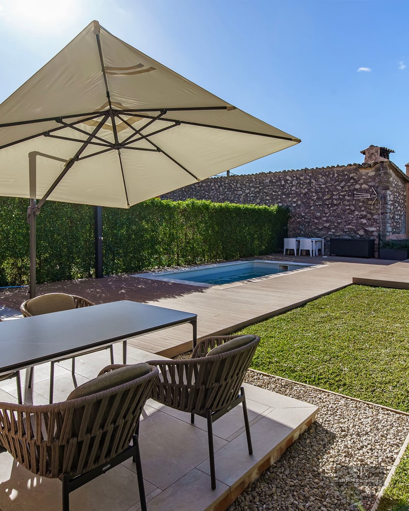 Sunlit terrace with modern dining set, large umbrella, small swimming pool, and lawn area with an olive tree.