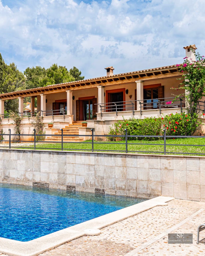Large blue swimming pool and sun loungers in front of a Mediterranean villa with a stone wall, lawn, and covered terrace.