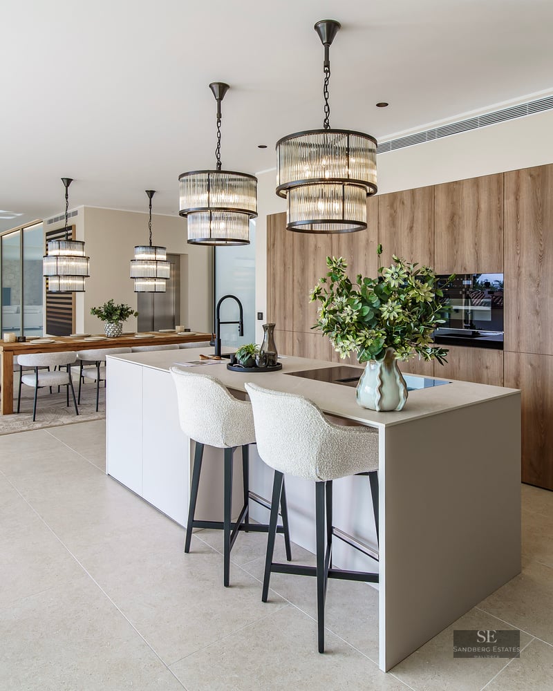 Modern kitchen with a large island, white stools, designer pendant lights, and wood cabinetry in an open-plan space.