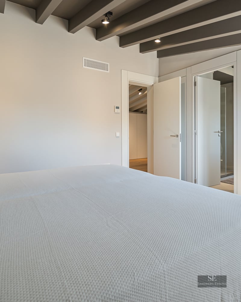 Bright bedroom featuring exposed ceiling beams, white bedding, built-in wardrobes, and light wood flooring.